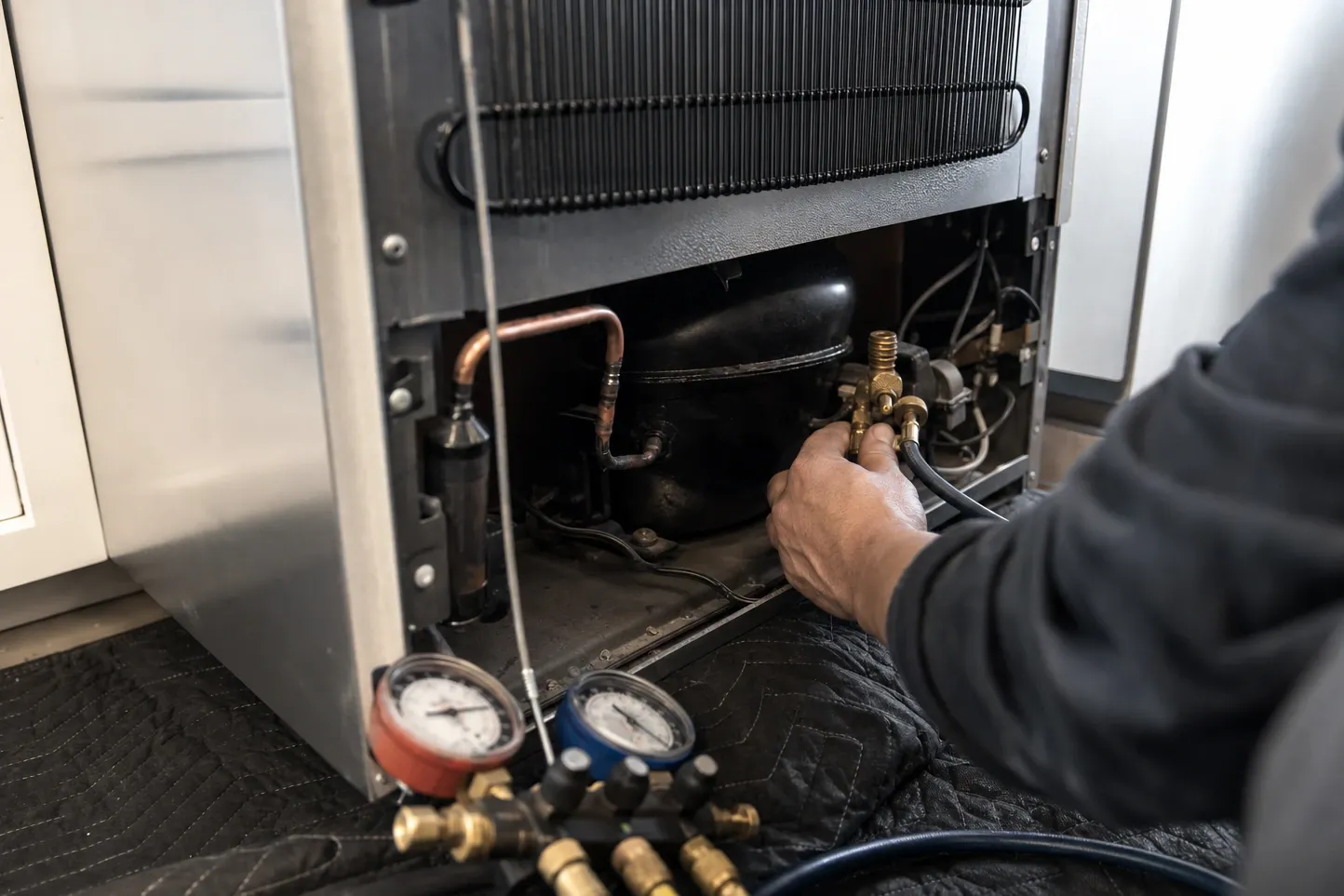 A hand fitting a hose to the brass service valve behind a refrigerator, condenser coil above the compressor and manifold gauges on a quilted blanket