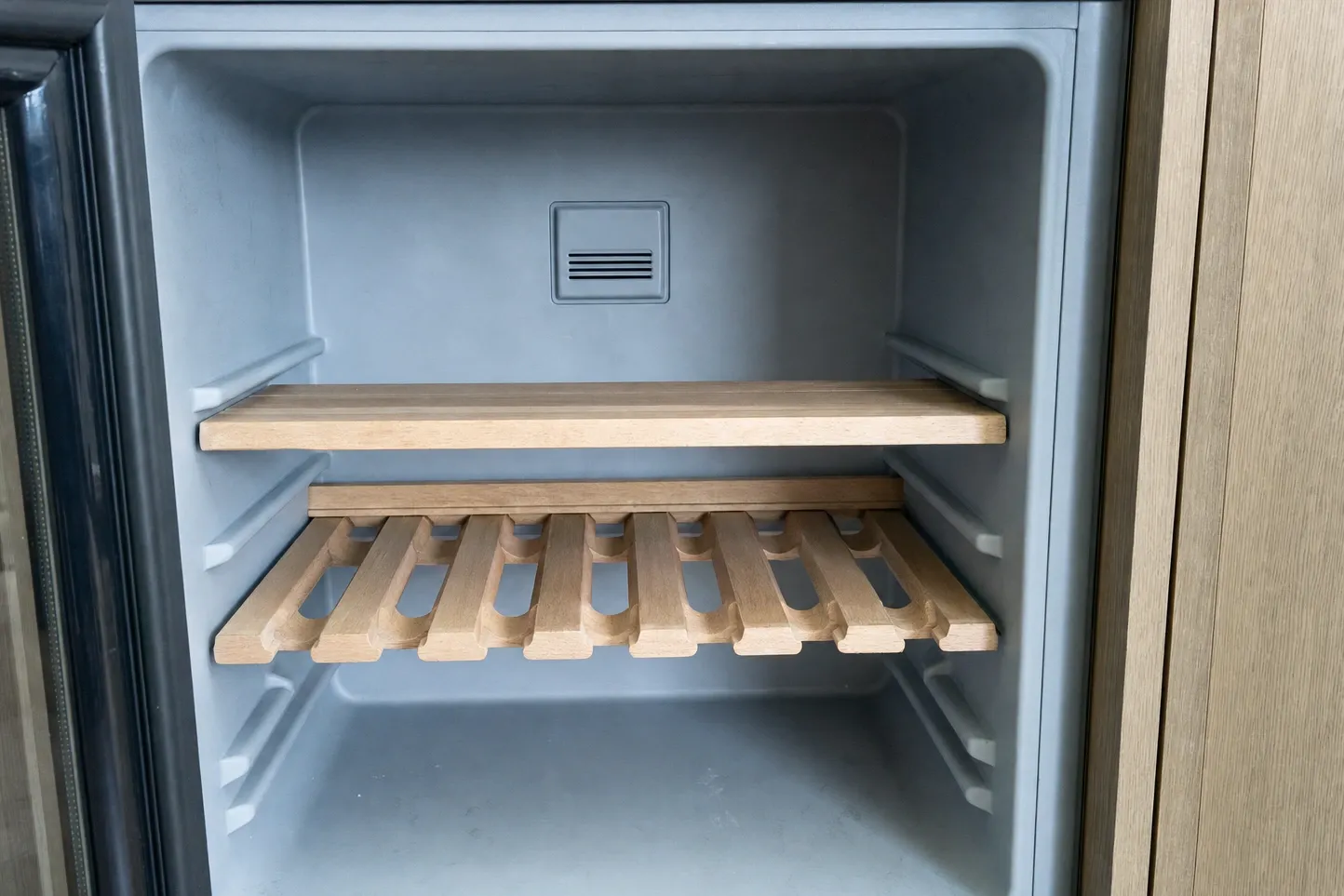 Empty interior of an undercounter wine cabinet: a solid wooden shelf above a slatted bottle rack, a vent grille in the back wall and the glass door open