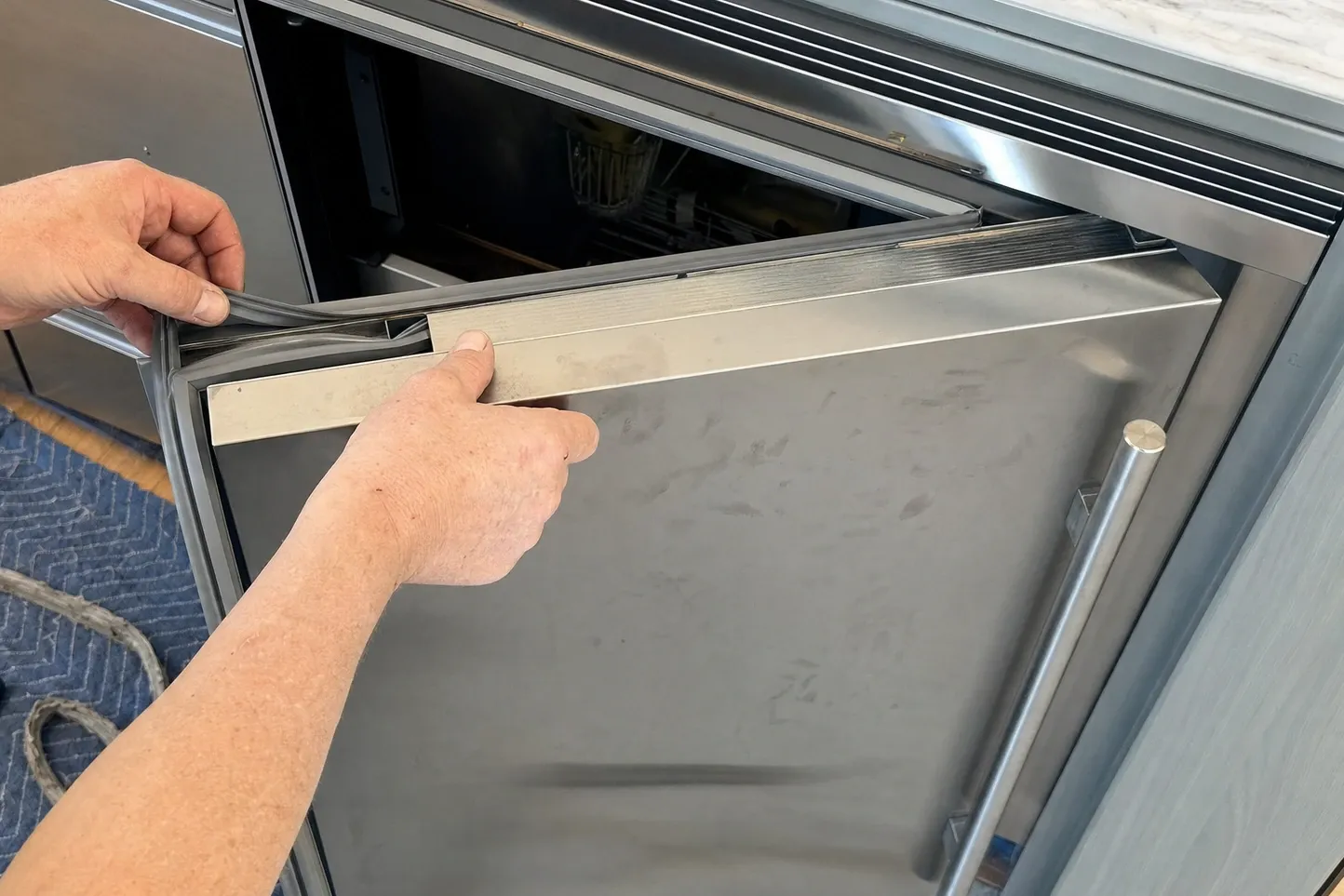 A hand peeling the grey gasket back from the inner top edge of an open stainless undercounter door while the other hand steadies the door trim rail