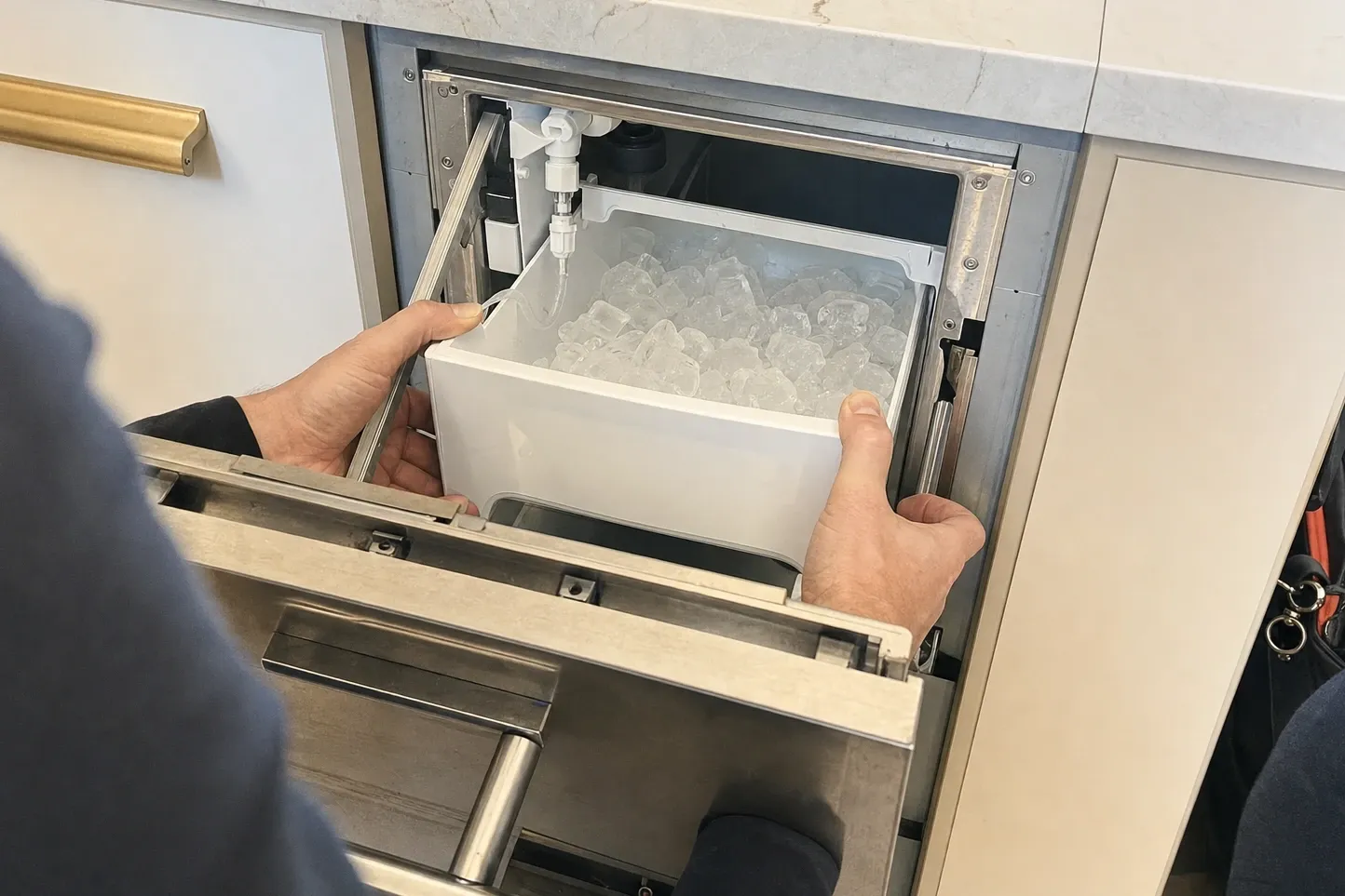 Hands lifting a bin heaped with clear cubes out of an undercounter ice drawer, the white fill spout and its tube hanging above the bin at the back
